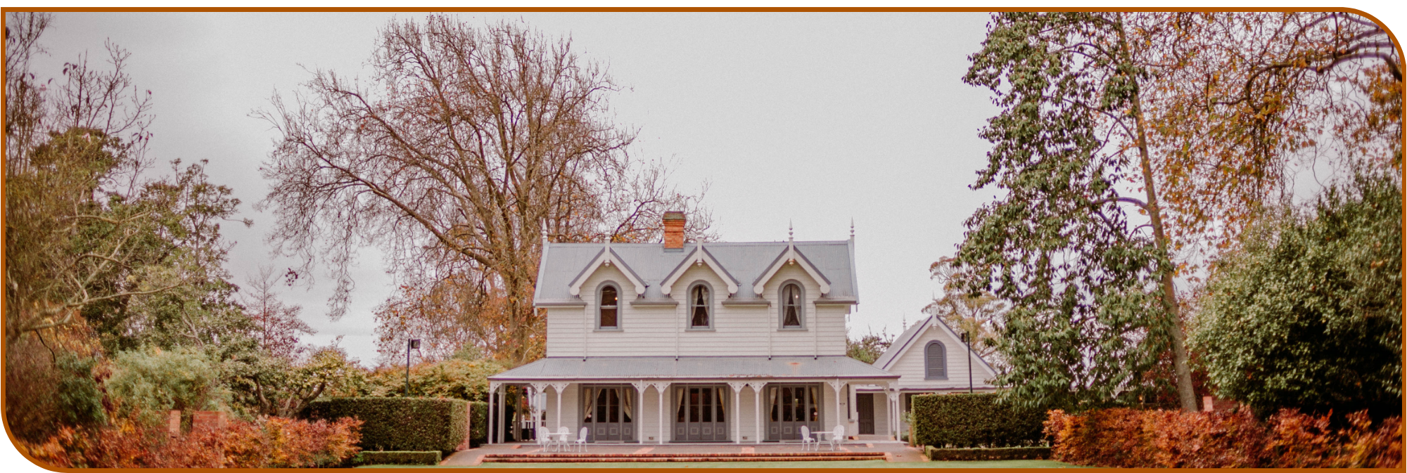 White Victorian-era homestead with steep roof, dormer windows and wrap-around veranda in formal hedged gardens with autumn trees