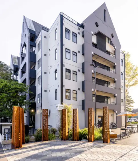 White and grey multi-storey building with angular roofline, rust-patinated carved columns at entrance