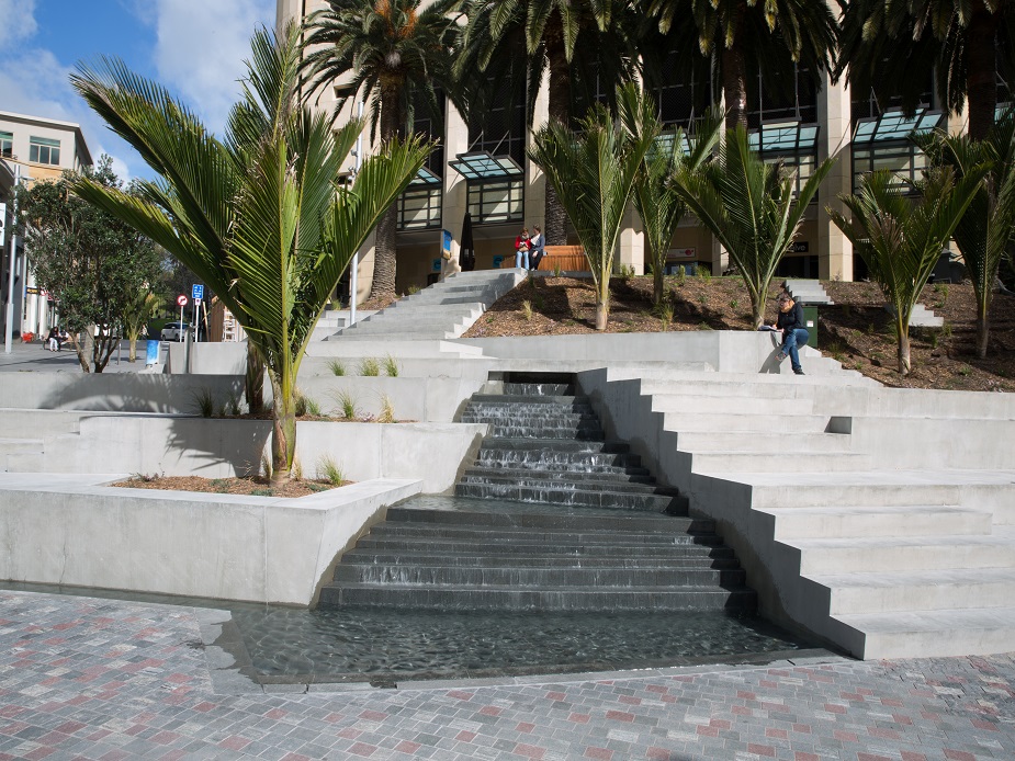 Water cascade over dark stone steps with trees and city buildings in background