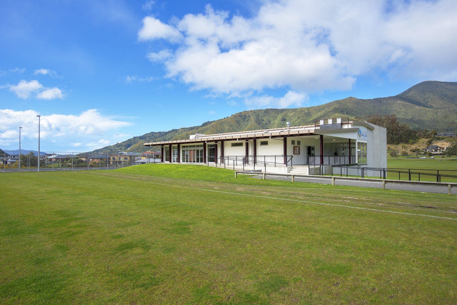 Single-storey pavilion with covered verandah, open to green sports fields and hills beyond