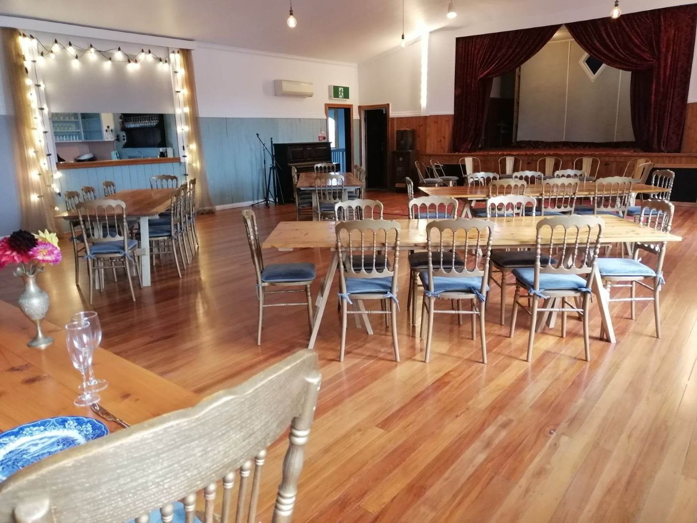 Hall interior with timber floor, dining tables with gold chairs, a bar to the left and a raised stage with red curtain