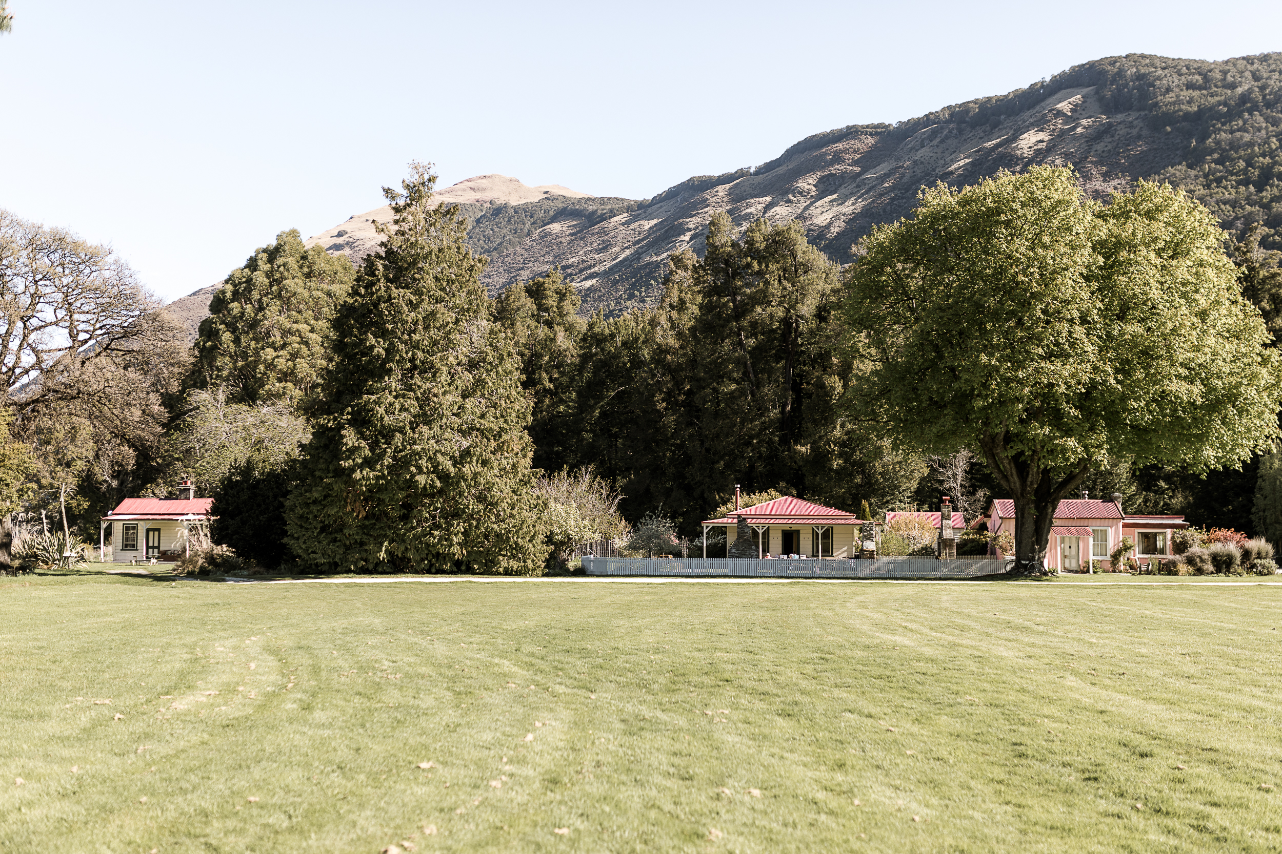 Red-roofed accommodation buildings across a wide flat lawn with mature trees and mountains behind