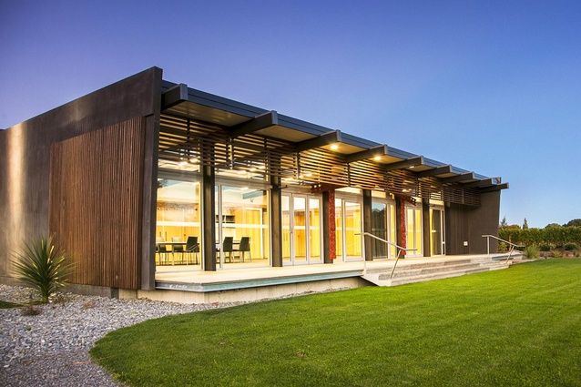 Low dark-clad building with timber louvred pergola, lit interior visible through full-height glazing, lawn foreground at dusk