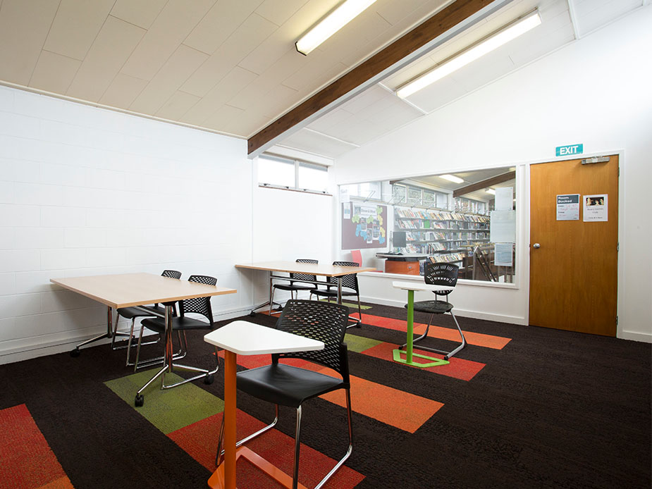Meeting room with tables, chairs, noticeboard and view into library through window