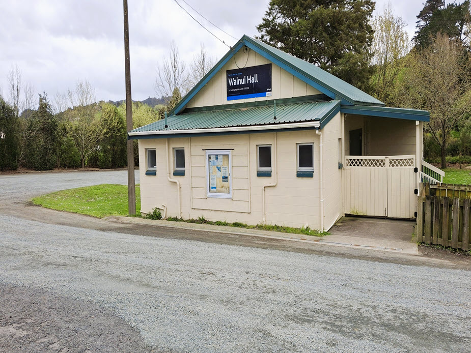 Small cream weatherboard hall with green metal roof beside a gravel road