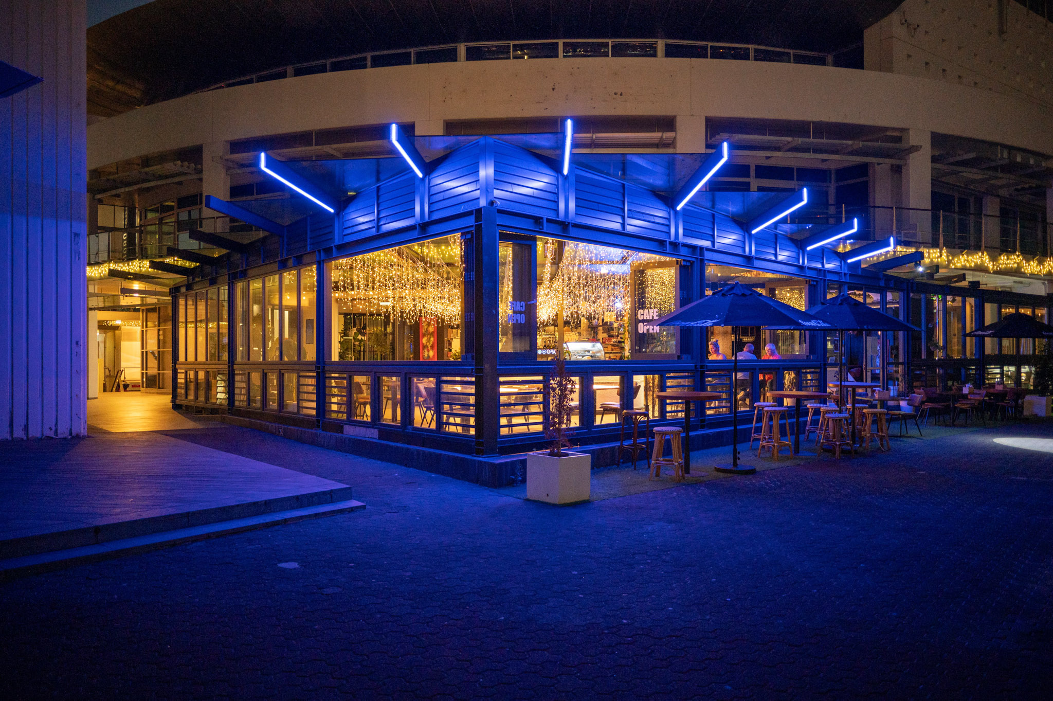 Glass-fronted venue with blue neon canopy, fairy lights inside, outdoor stools at night