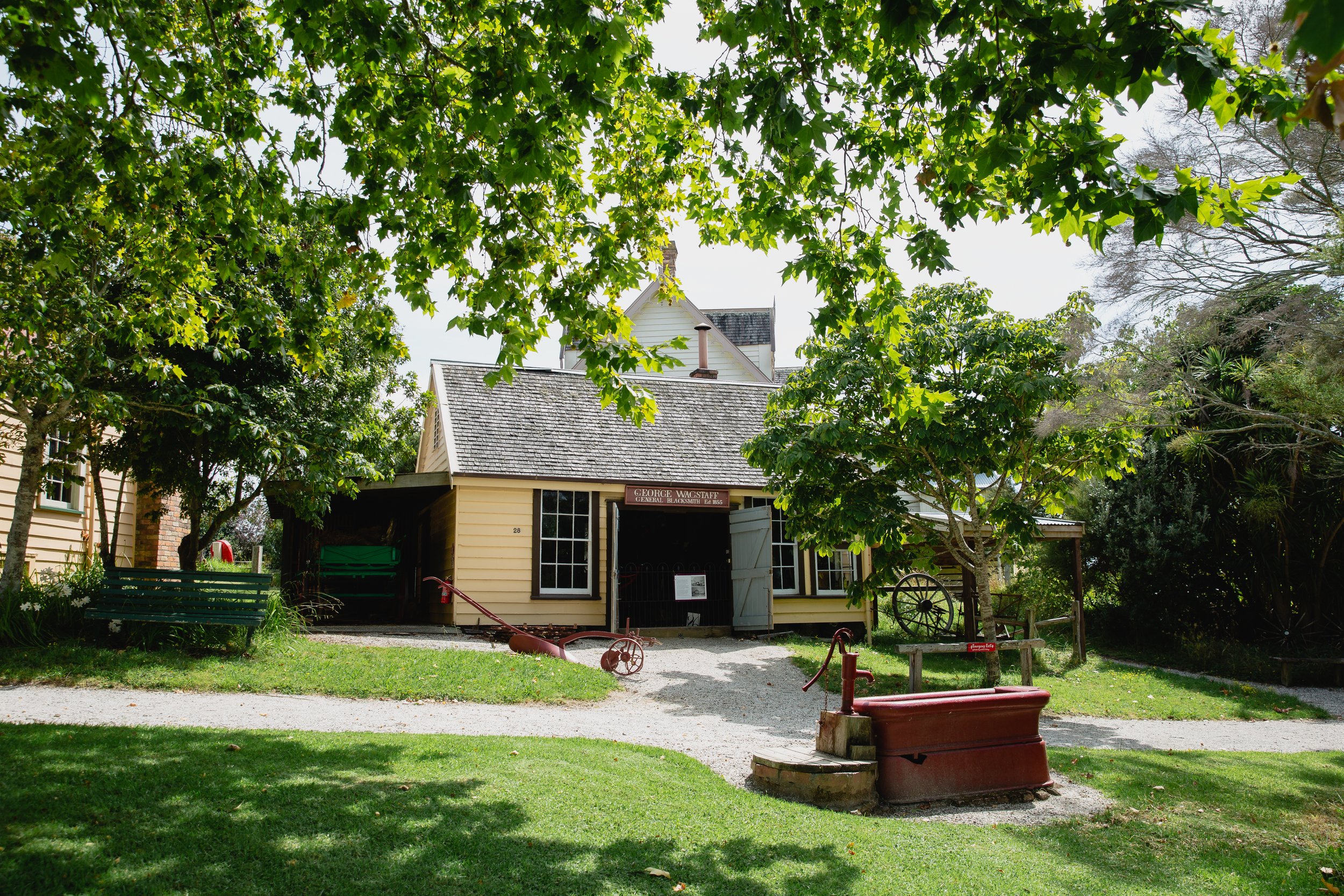 Yellow weatherboard colonial buildings with gravel courtyard, trees and red farm implements