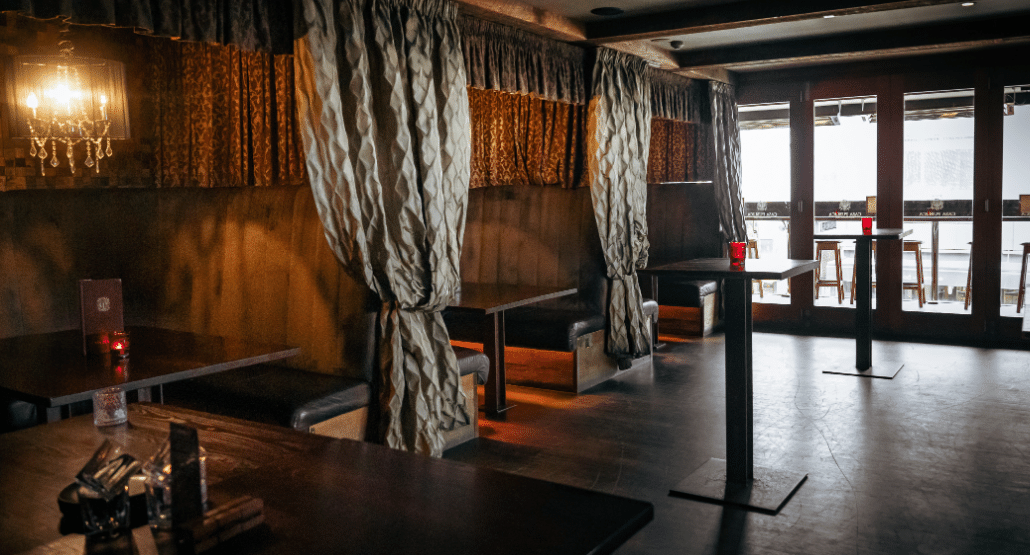 Darkened bar interior with draped fabric booth dividers, chandelier, and high stools along tall tables