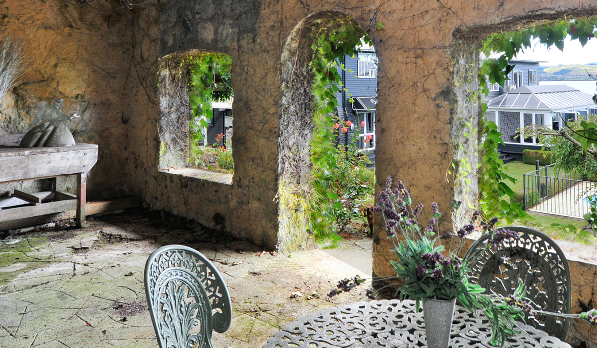 Weathered stone garden wall with open rectangular windows draped in climbing plants, cast-iron furniture on stone floor
