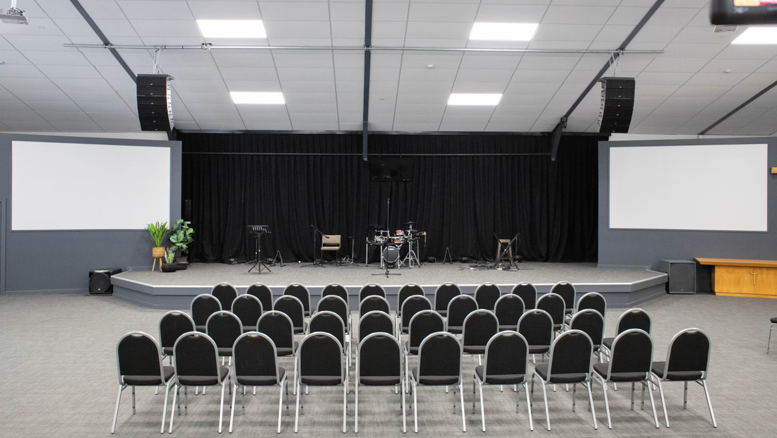 Modern church hall with rows of chairs facing a raised platform, flanked by large screens and a lighting rig