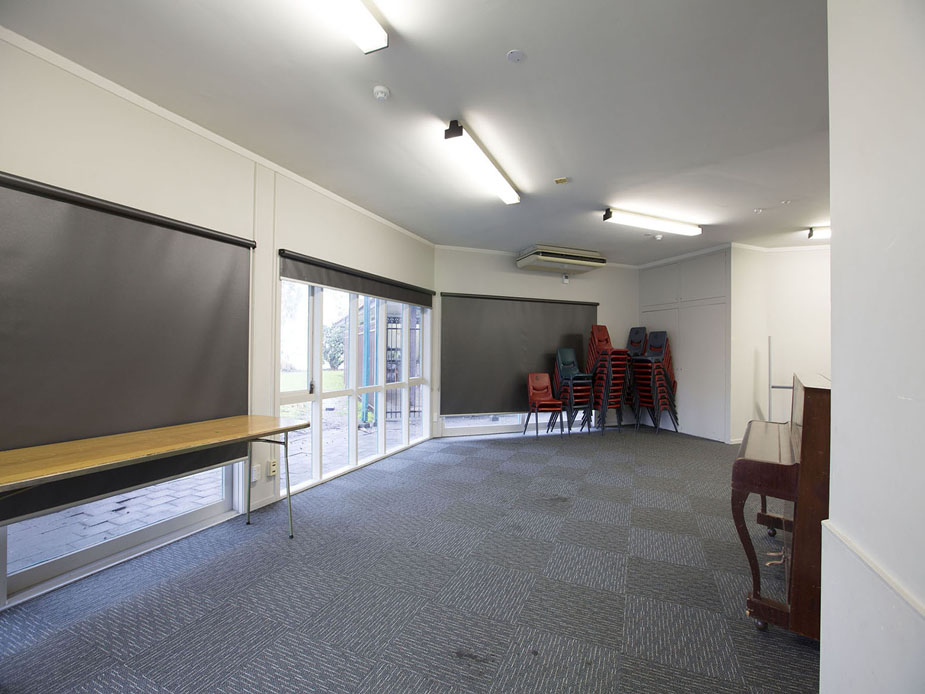 Plain carpeted room with stacked chairs, roller blinds, and upright piano