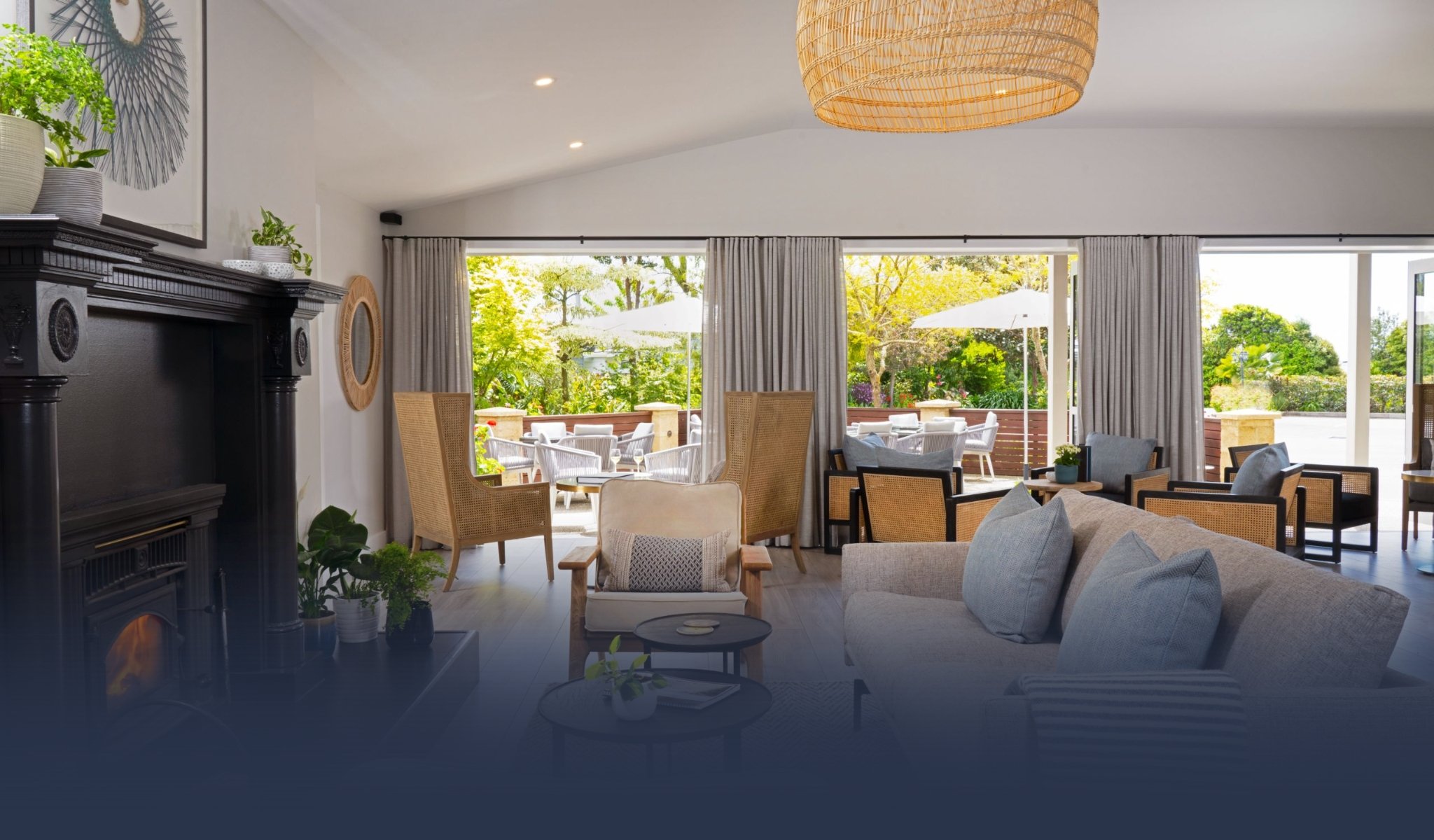 Lounge interior with fireplace, neutral sofas, woven pendant light, and open bifold doors to a terrace