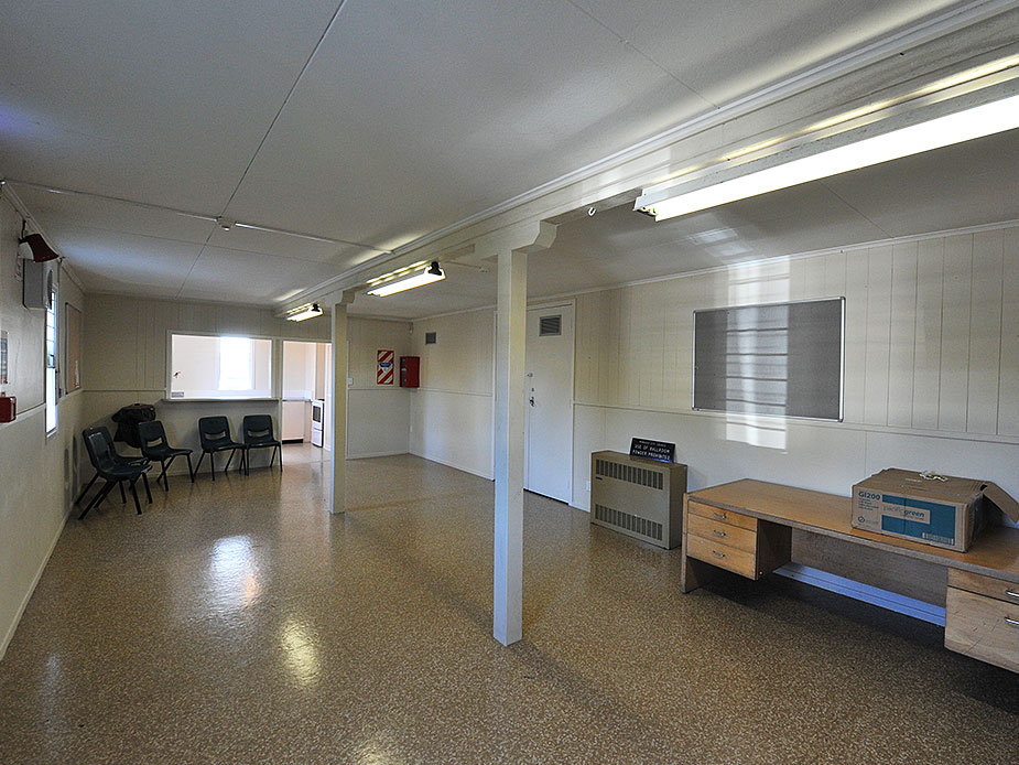 Plain community hall with low ceiling, a few chairs, and servery hatch