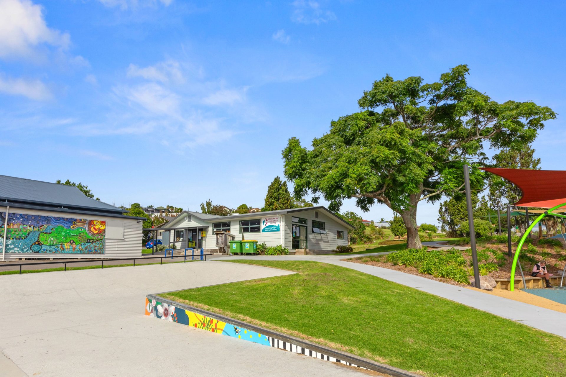 Single-storey community building and playground area with trees and shade sail