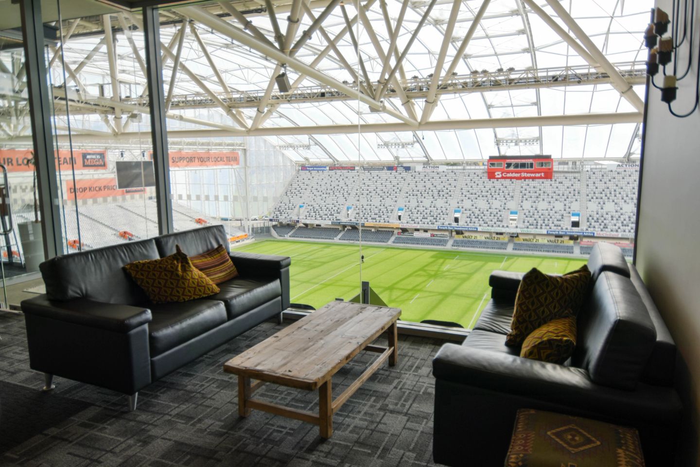Stadium lounge with black leather sofas, timber coffee table, floor-to-ceiling windows overlooking playing field