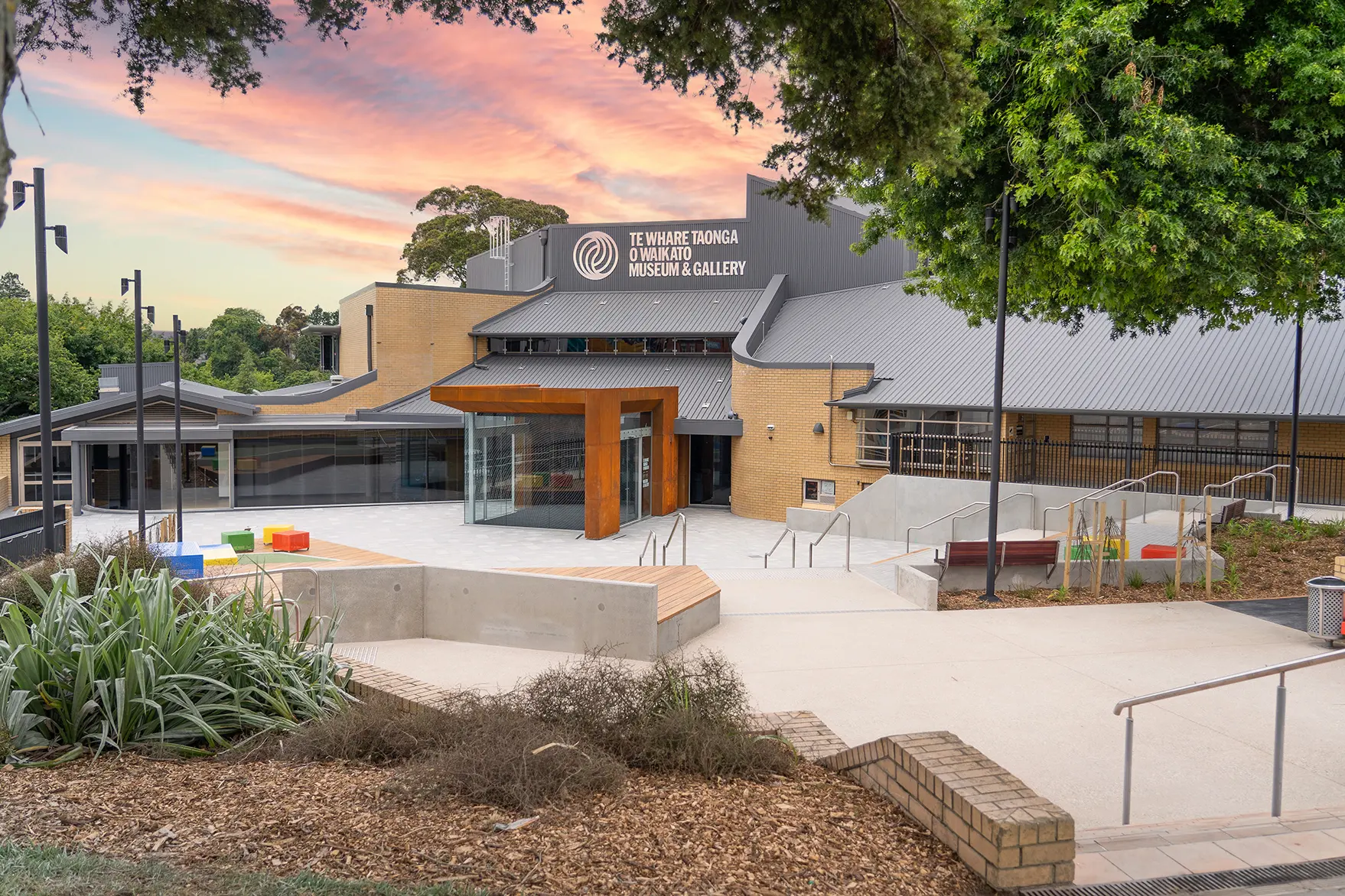 Brick and metal-roof building with timber-framed glazed entry, concrete forecourt, gardens at dusk