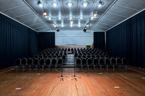 Hall interior with rows of chairs facing a draped stage lit in blue and purple