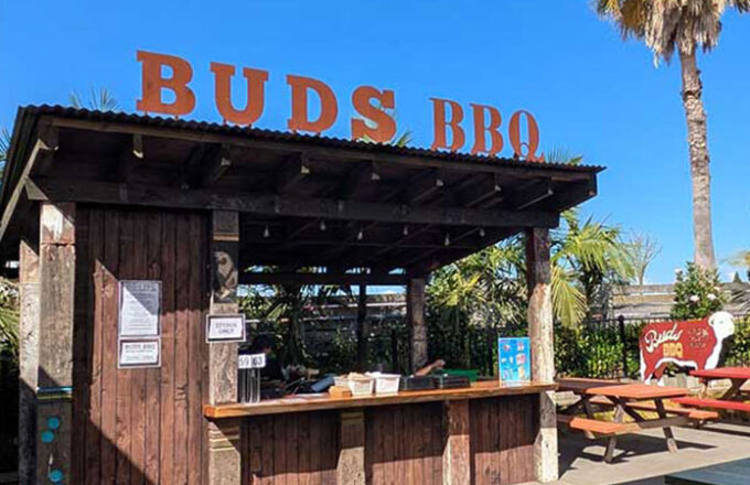 Outdoor rustic bar kiosk with Buds BBQ signage and picnic seating