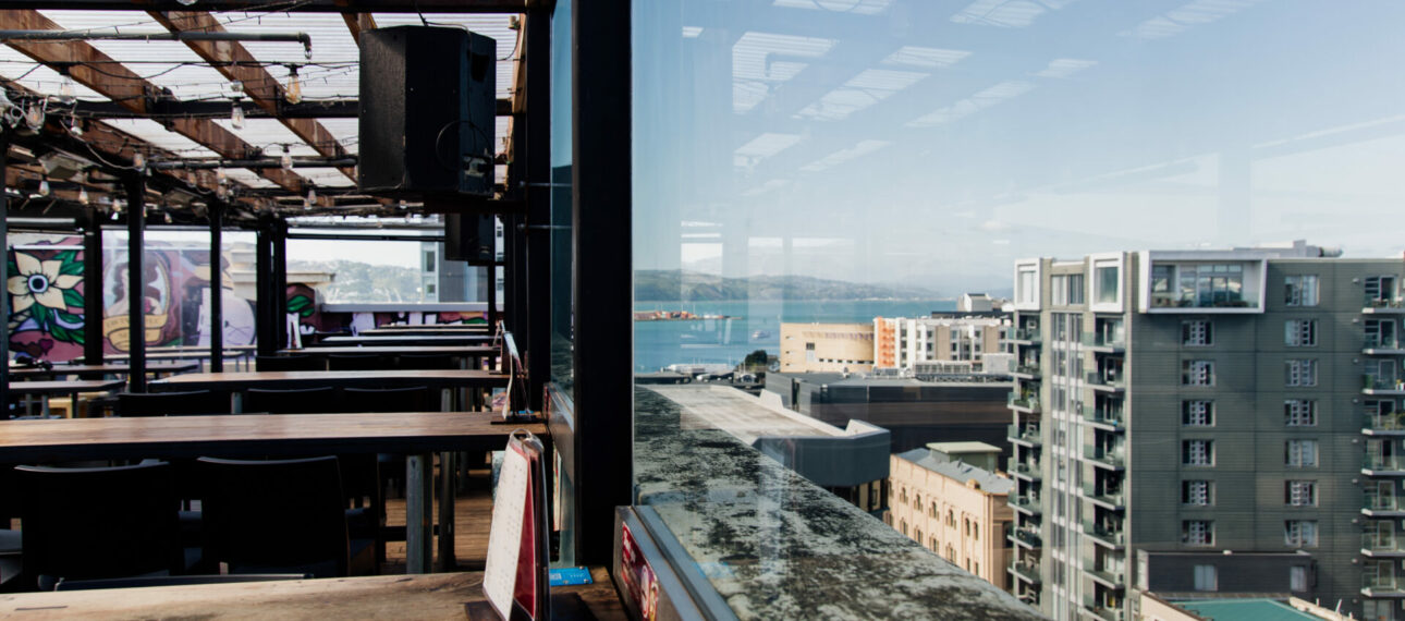 Open rooftop bar interior by day with exposed steel beams, long wooden benches, and large window overlooking city