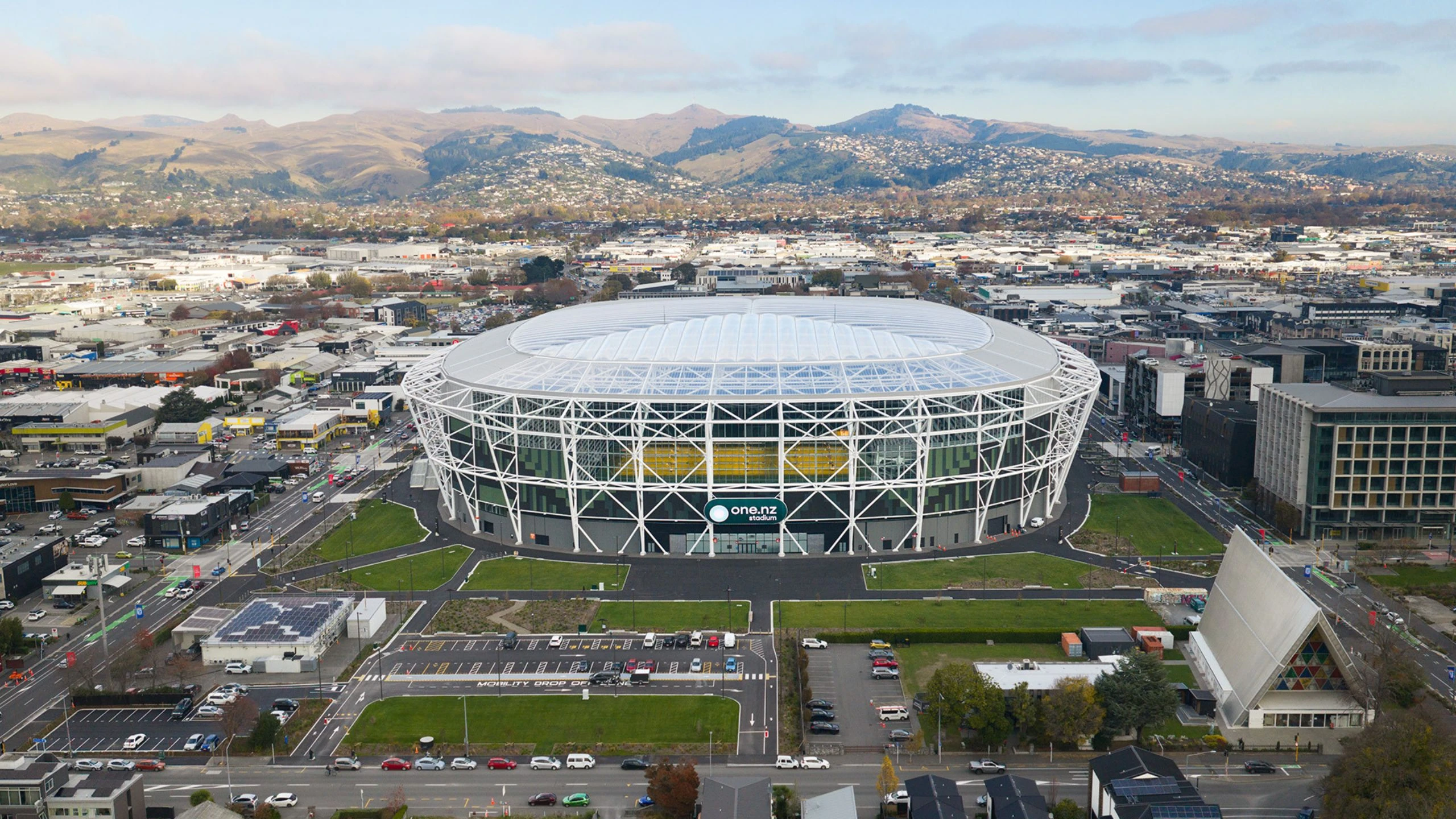 Large oval stadium with white lattice steel frame and translucent roof, aerial view in an urban setting with hills behind