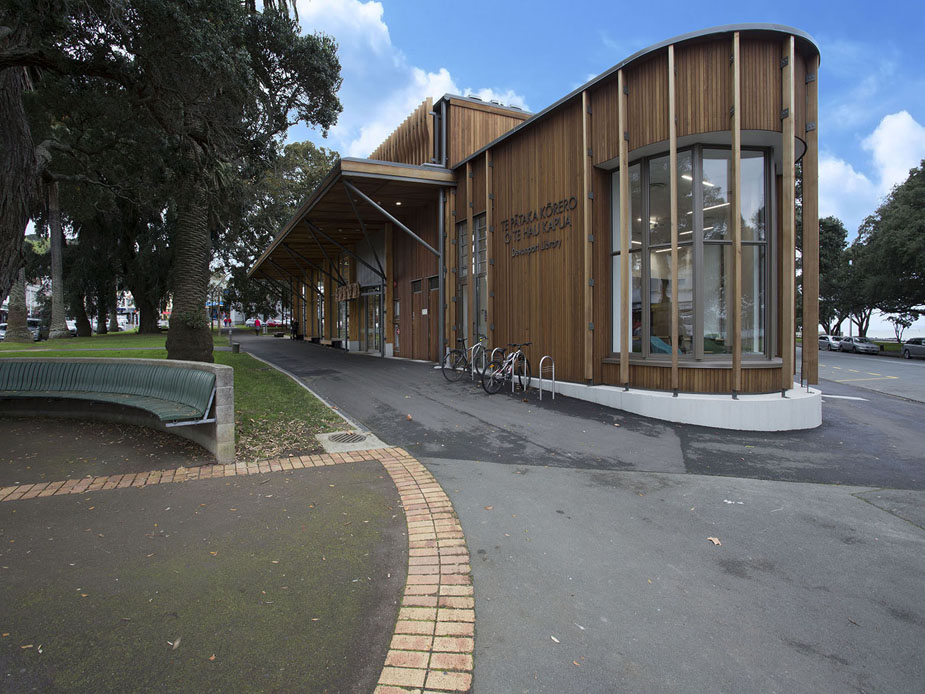 Contemporary timber-clad building with curved glass corner section, footpath and park bench in foreground