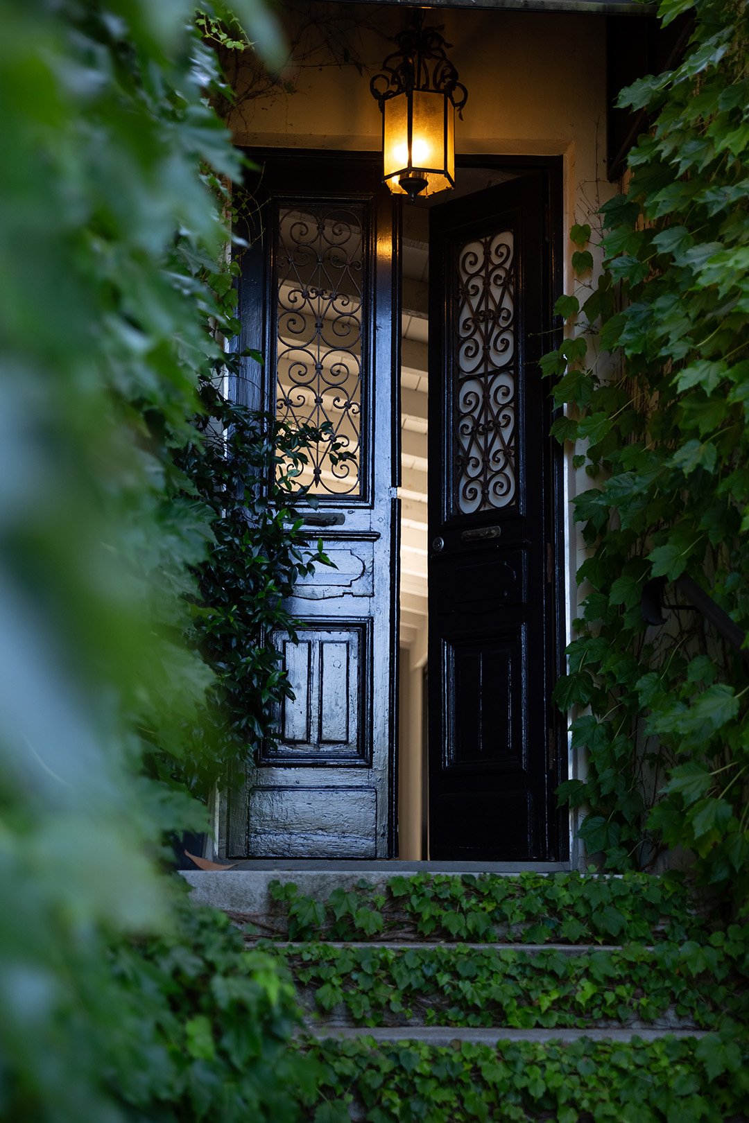Dark iron-wrought entrance doors with ornate scrollwork panels, ajar to reveal lit interior, stone steps and climbing plants