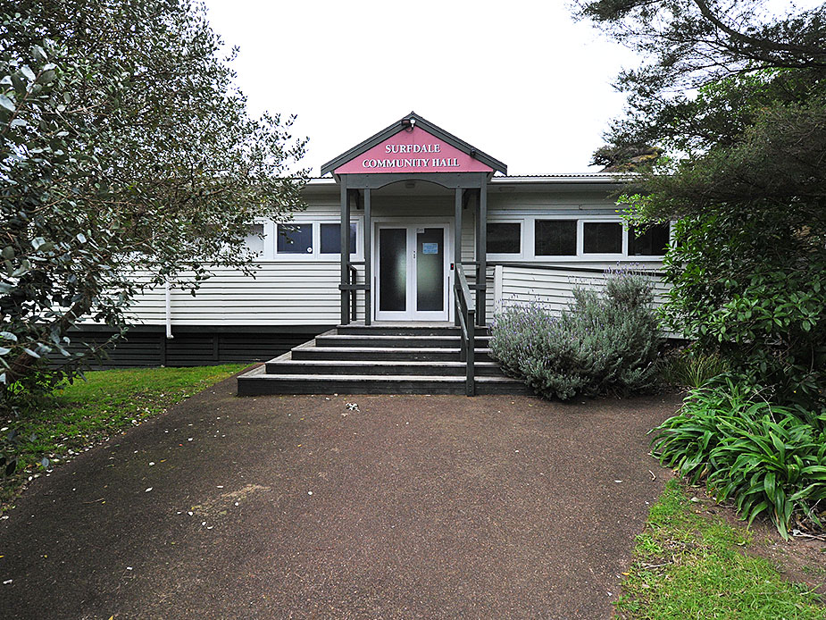White weatherboard hall with entry steps and pink fascia sign, surrounded by planting