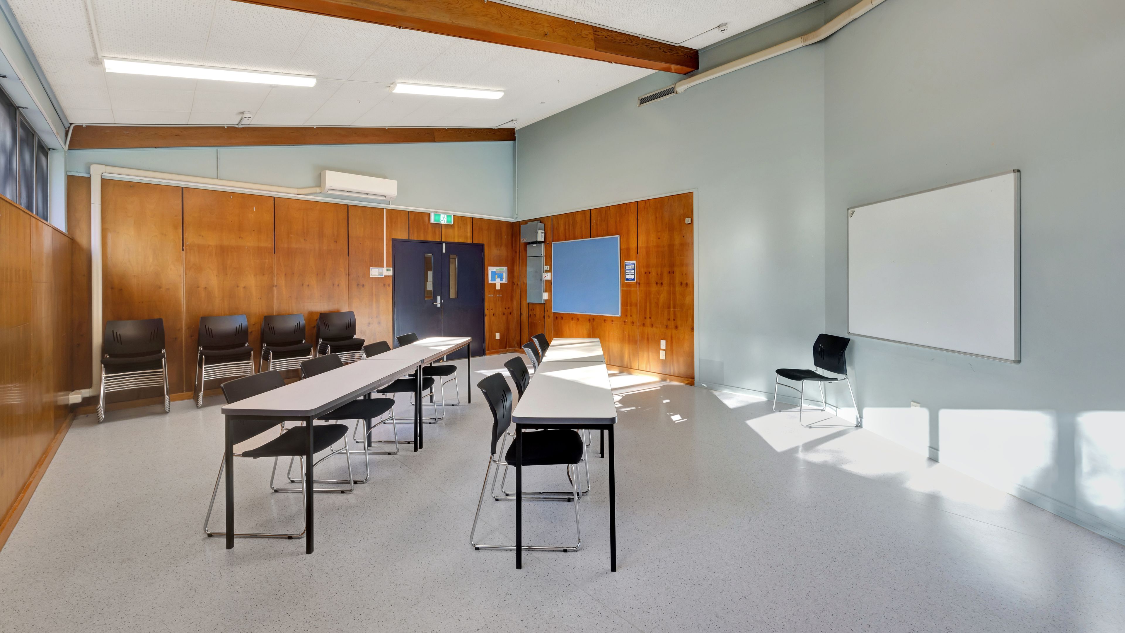 Small meeting room with folding tables, black chairs, timber-panelled wall, whiteboard, and large windows