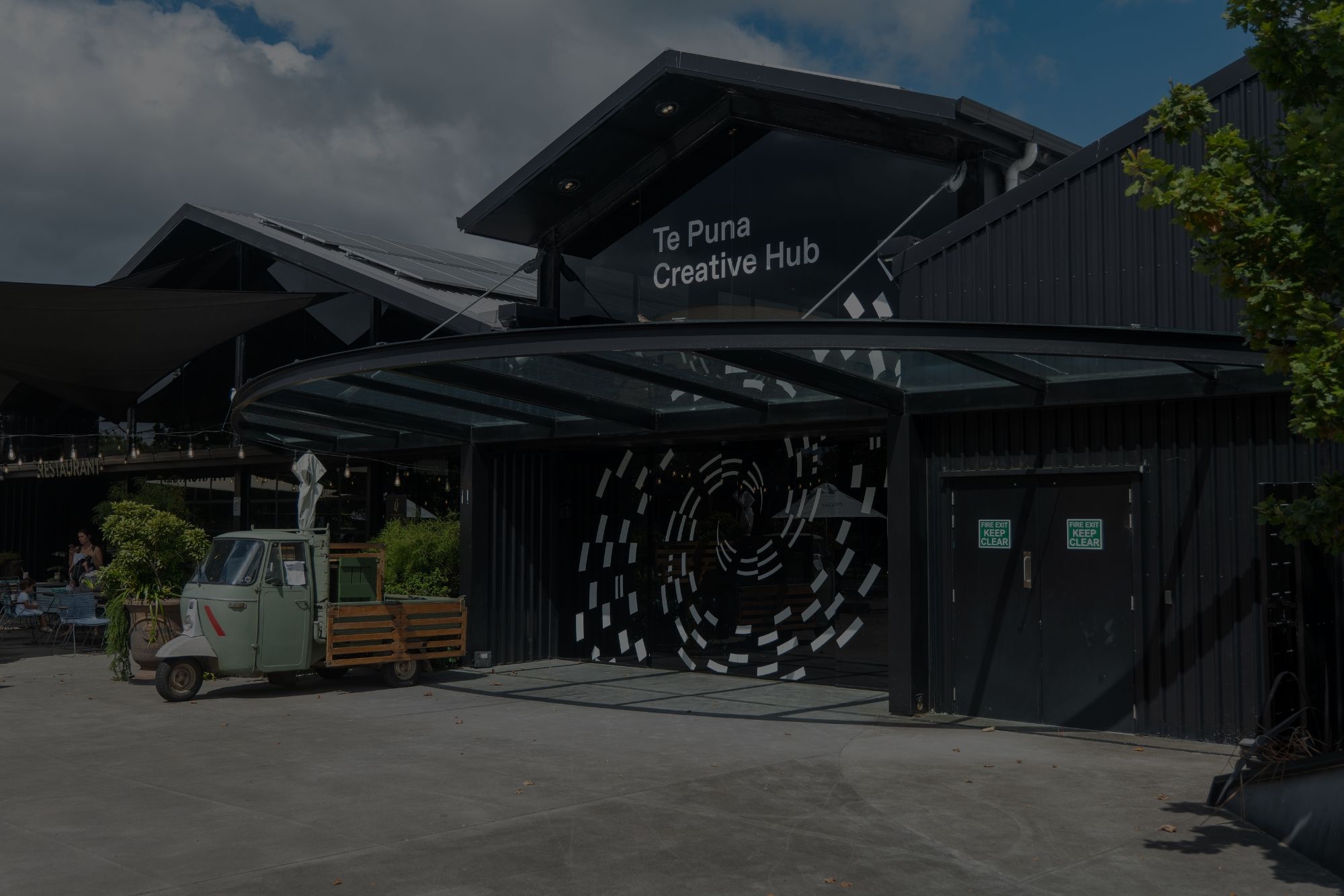 Dark timber-clad building with covered entry, large spiral mural on wall, outdoor courtyard