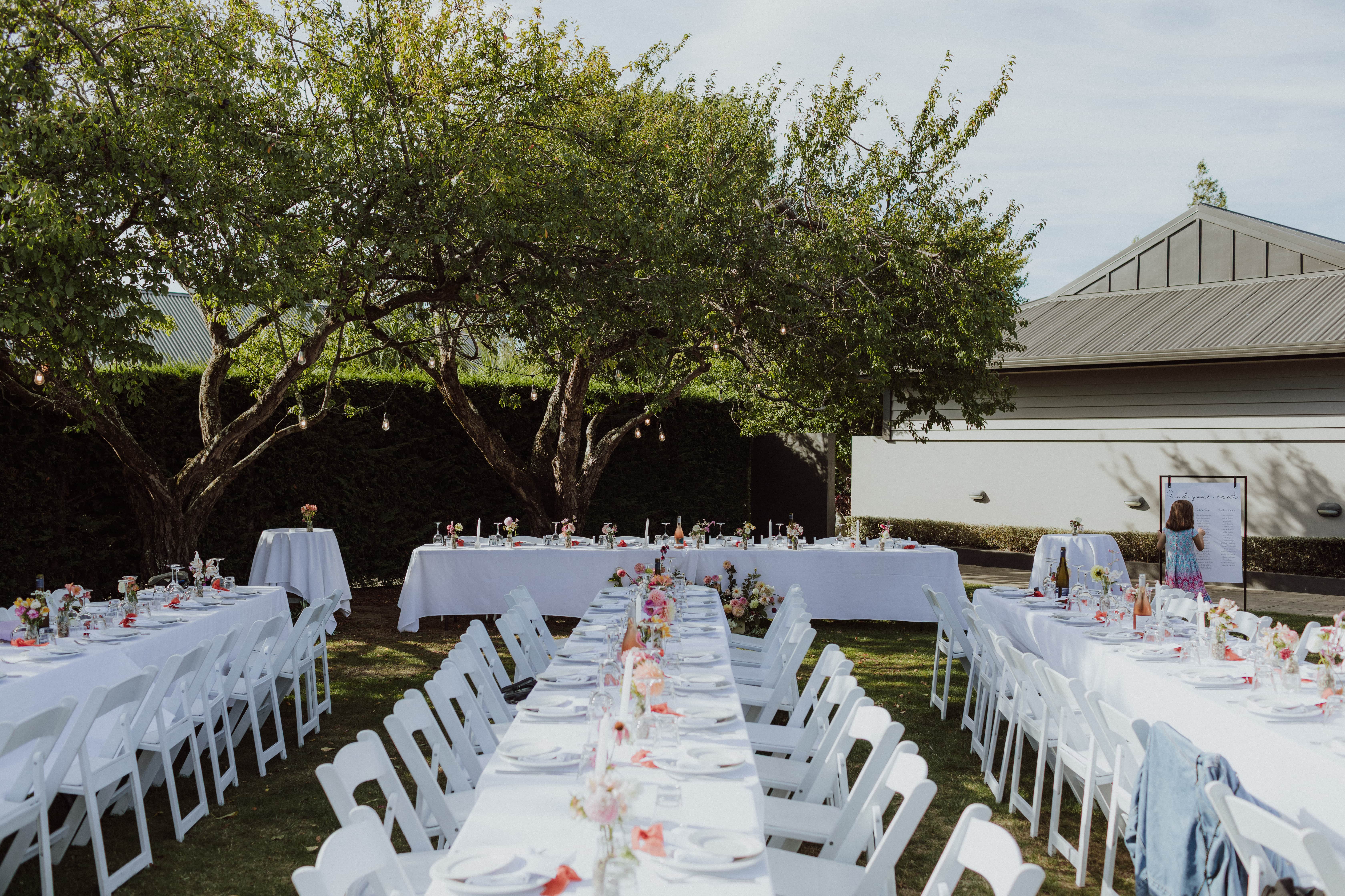 Outdoor reception with white linen tables, white folding chairs, festoon lights and mature trees
