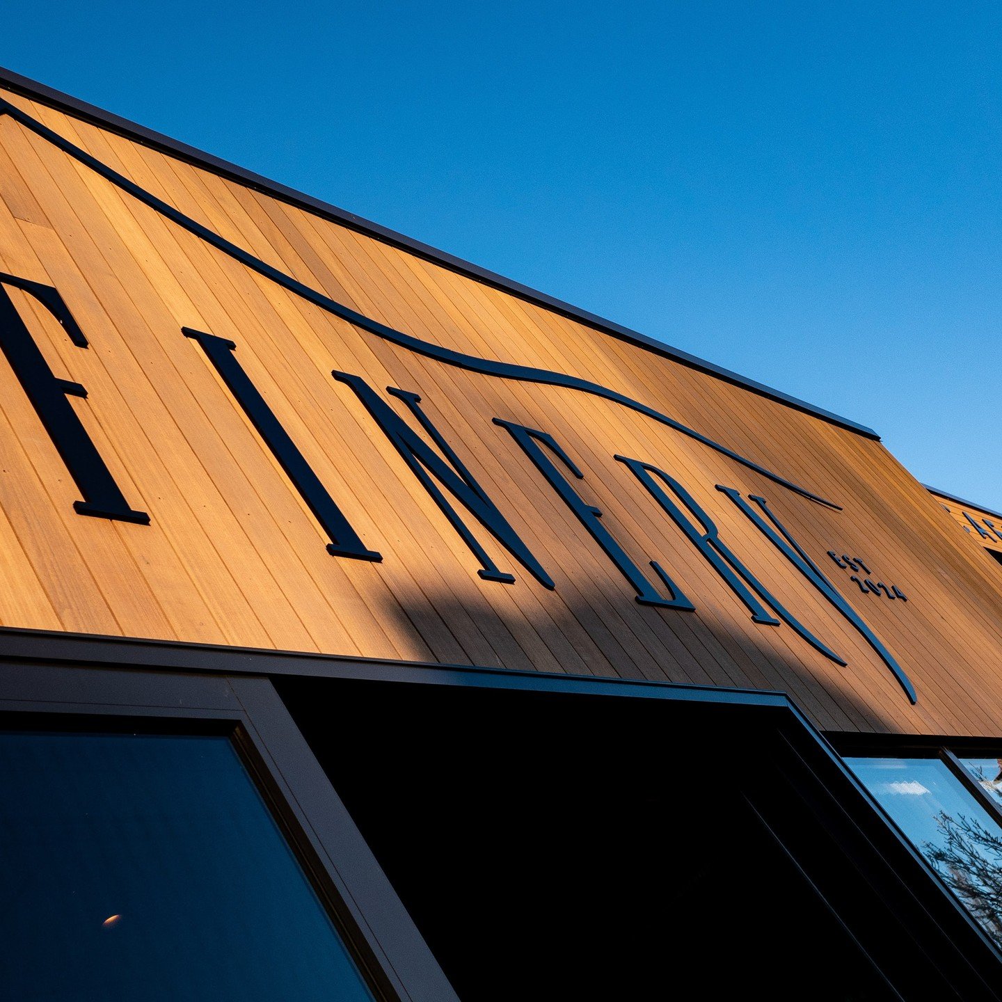 Warm timber-clad barn gable end with large cut-out signage, angular roof, clear blue sky
