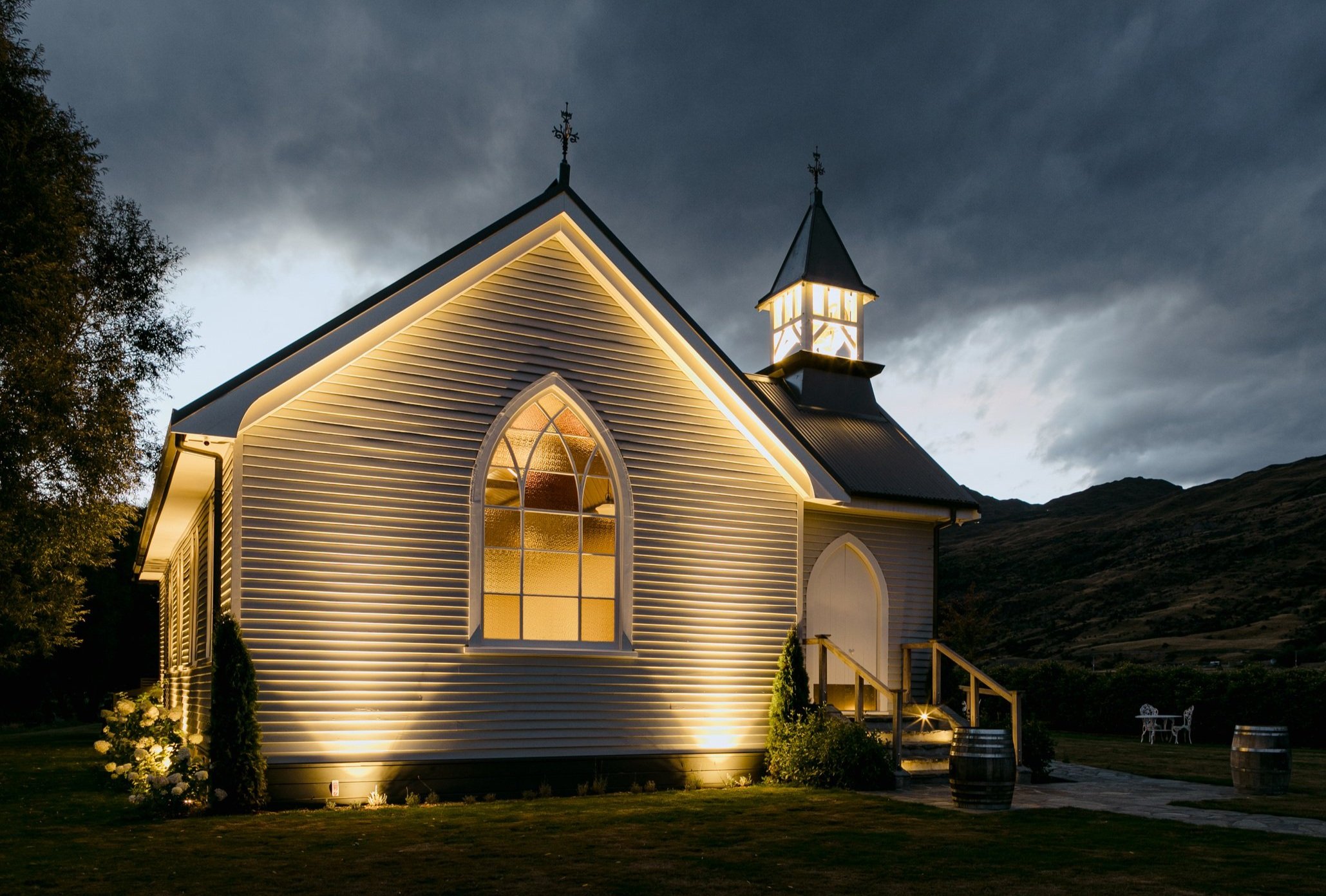 White timber church exterior lit at night with glowing arched window and dark hill beyond