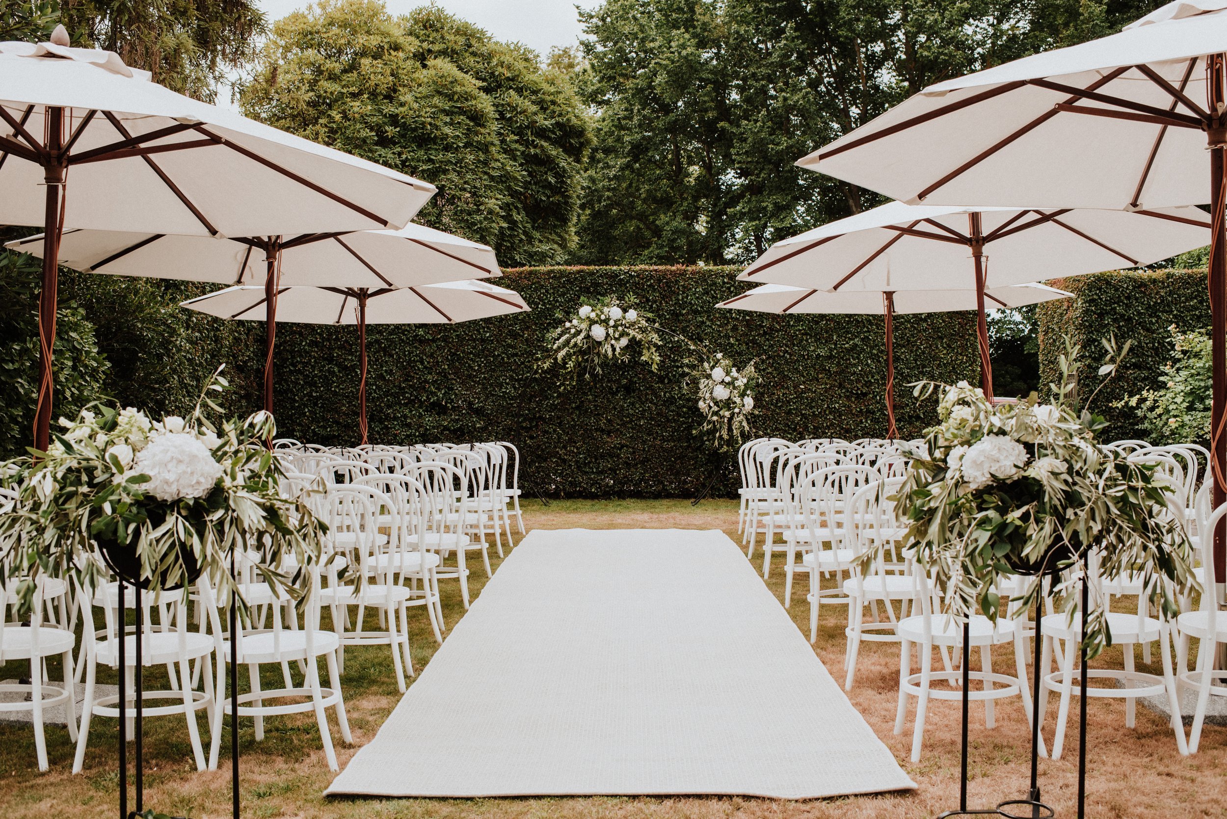 Outdoor ceremony on paved terrace with aisle, white chairs and trimmed hedgerow surrounds