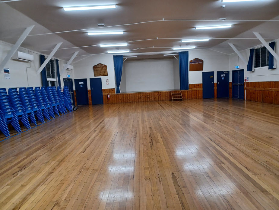Large open hall with polished timber floor, stacked chairs, and projection screen