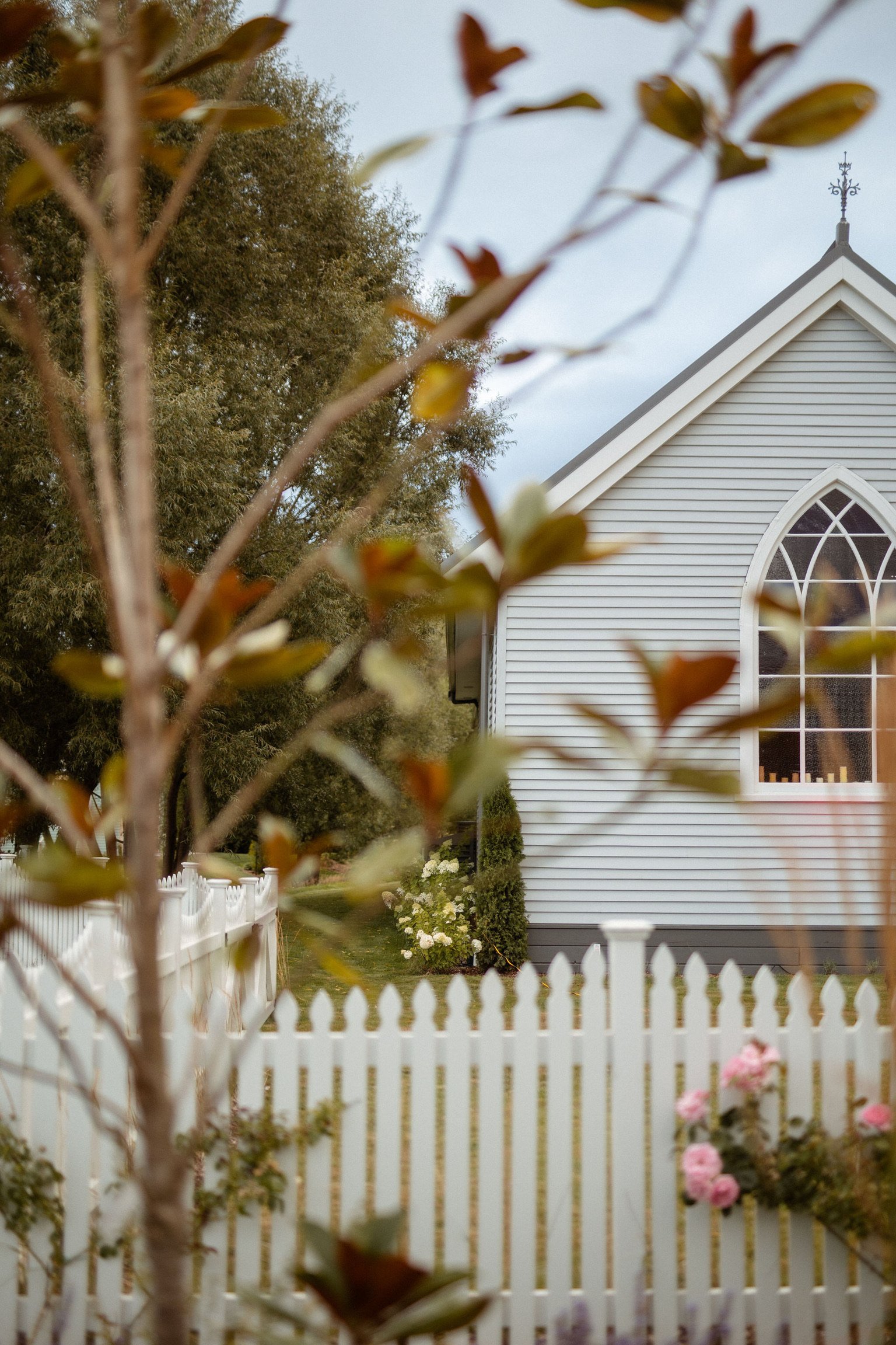 White timber church with Gothic arch window and white picket fence seen through trees