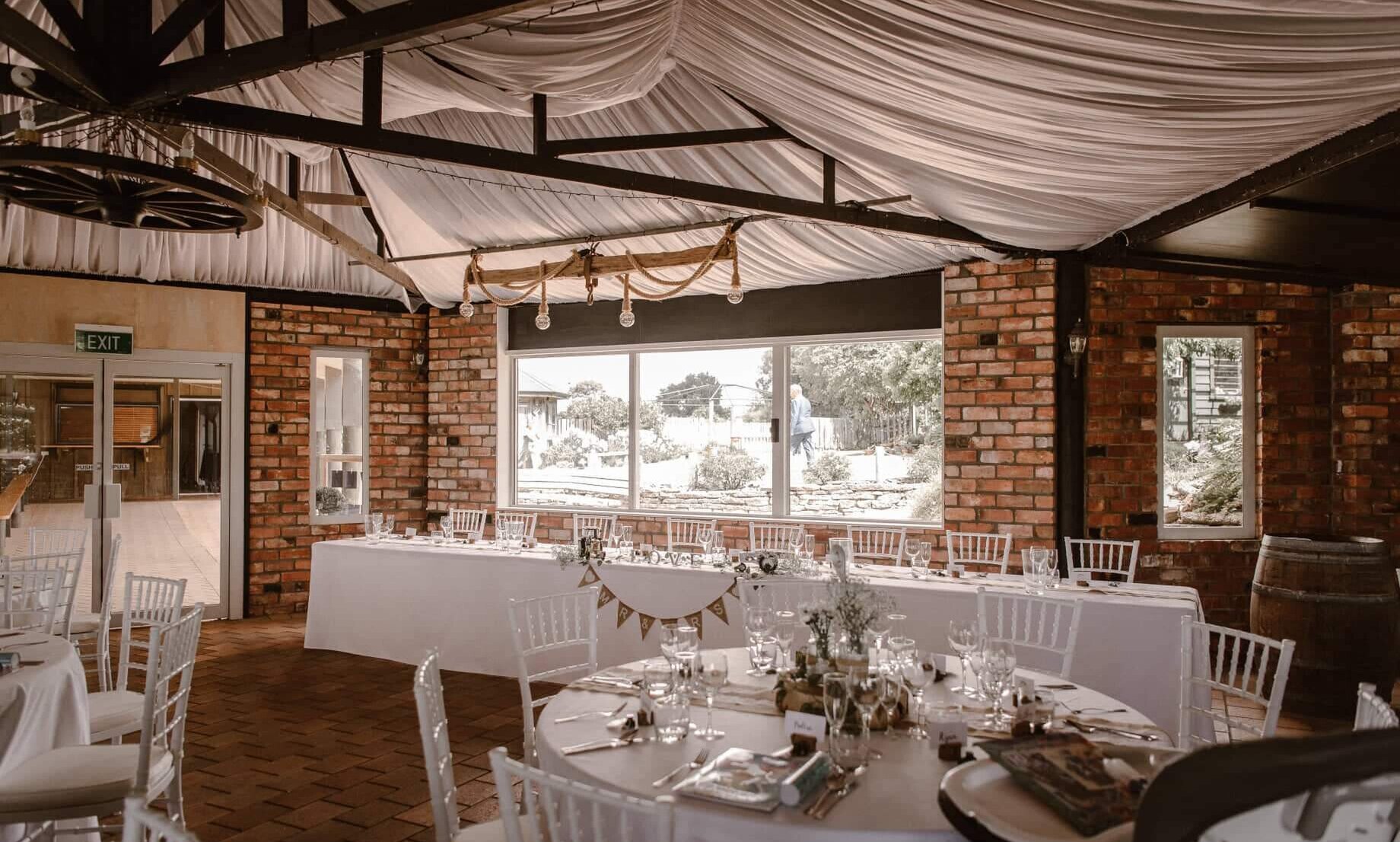 Brick-walled function room with exposed trusses, draped white fabric ceiling, round tables set for dining