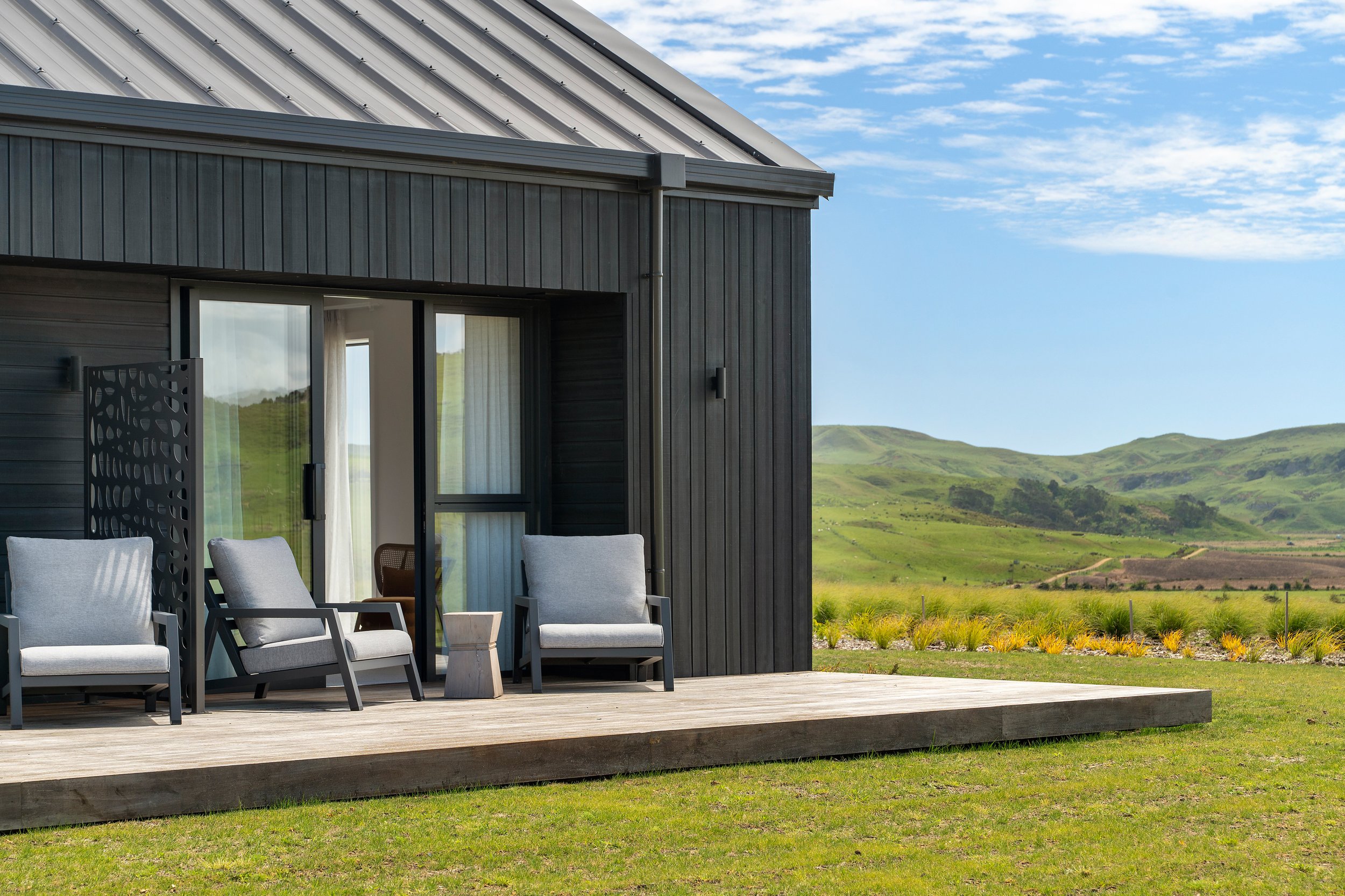 Dark vertical-clad building with timber deck, two outdoor chairs, and green hillside view