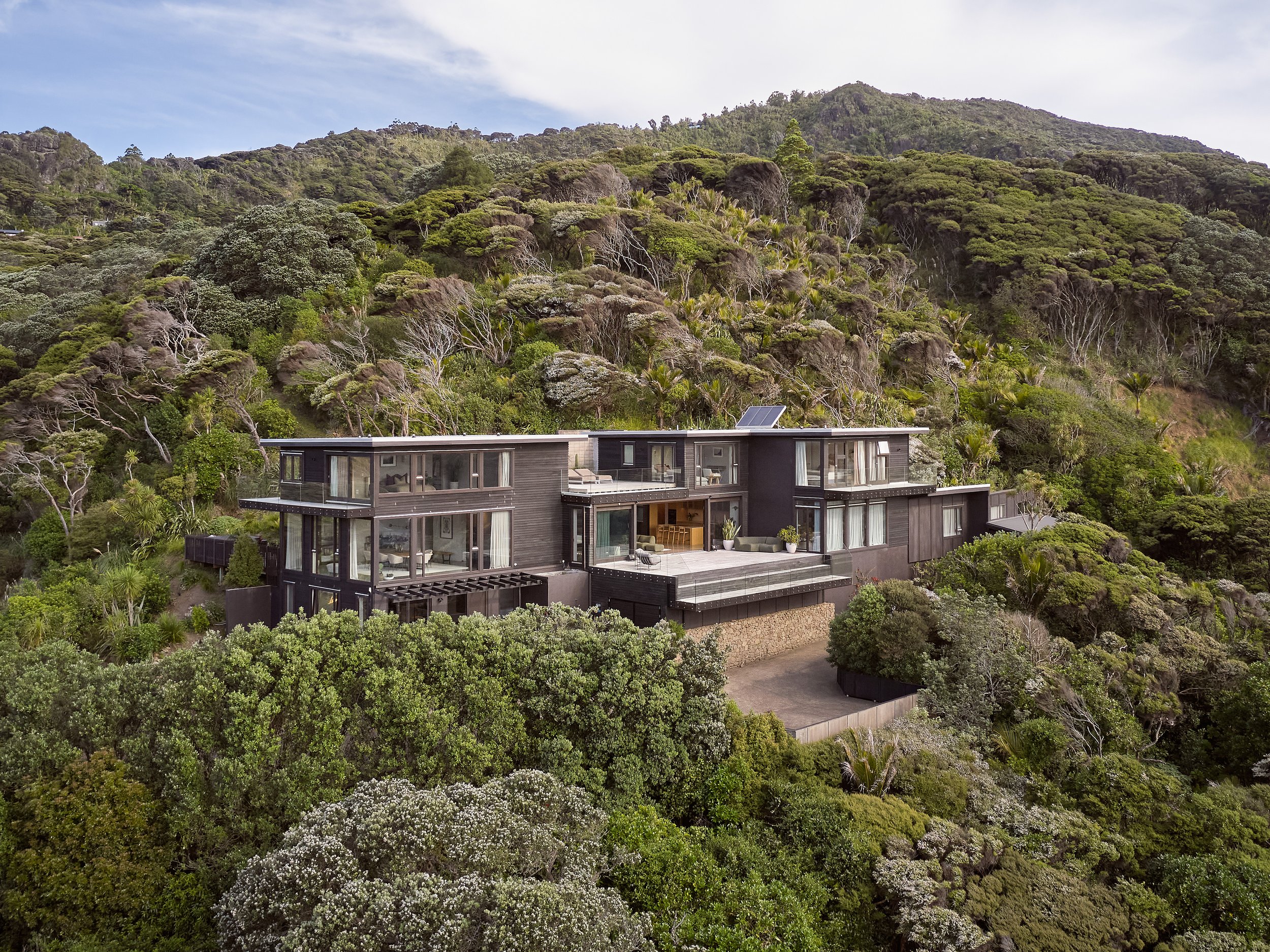 Dark-clad multi-level house with large windows set into dense bush-covered hillside