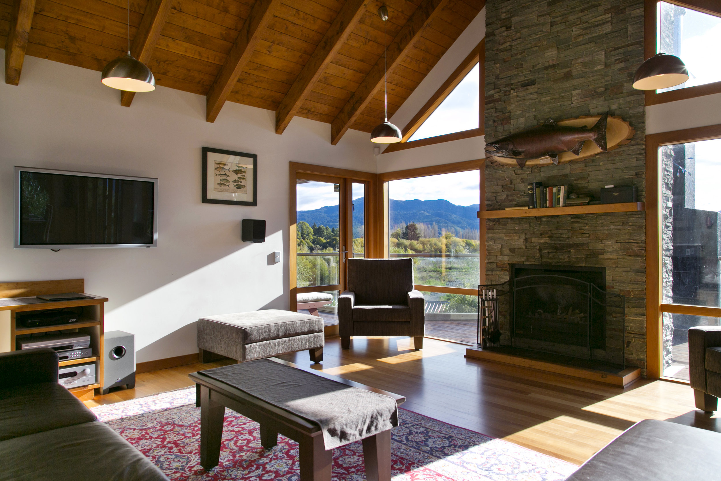 Open-plan lounge with exposed timber beams, stone fireplace, and floor-to-ceiling windows to mountains