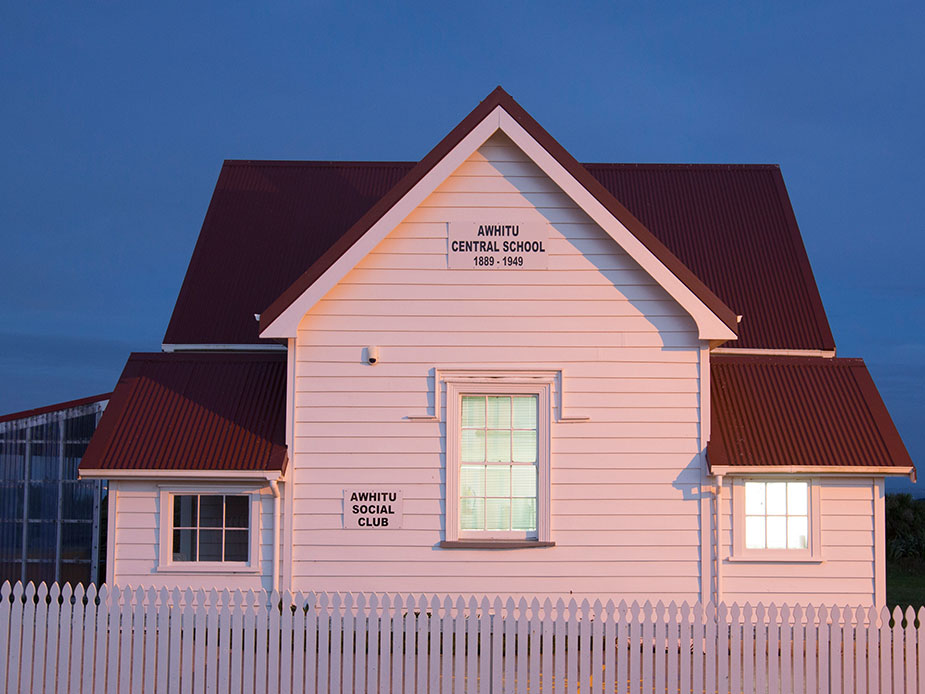 White weatherboard hall with red iron roof and picket fence, lit at dusk