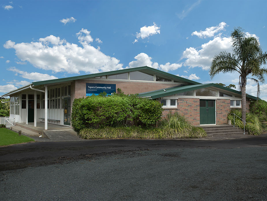 Low-set brick hall with green metal roof, covered verandah, and a palm tree alongside