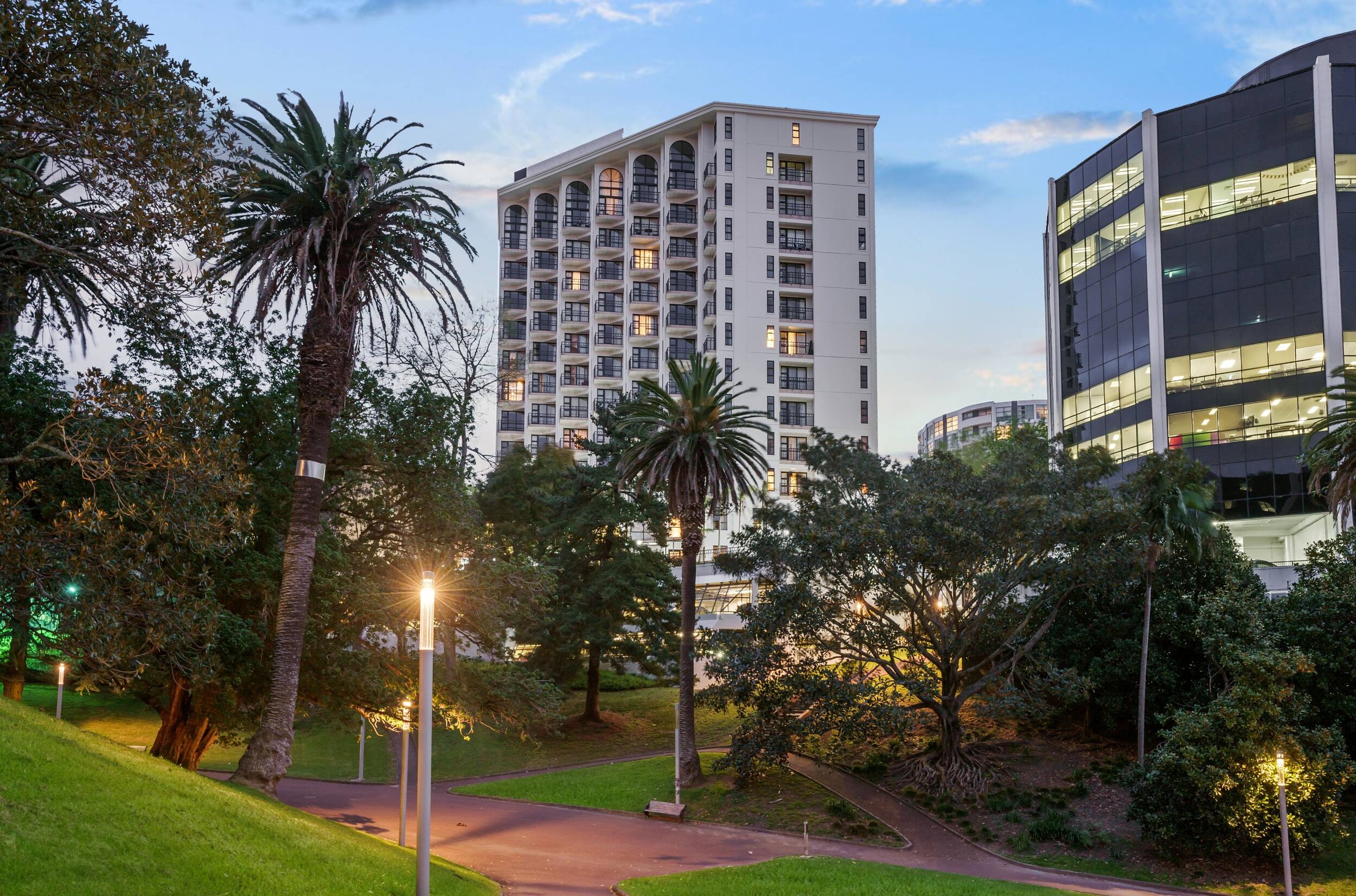 Tall white hotel tower with arched windows, seen from a park with tall trees at dusk