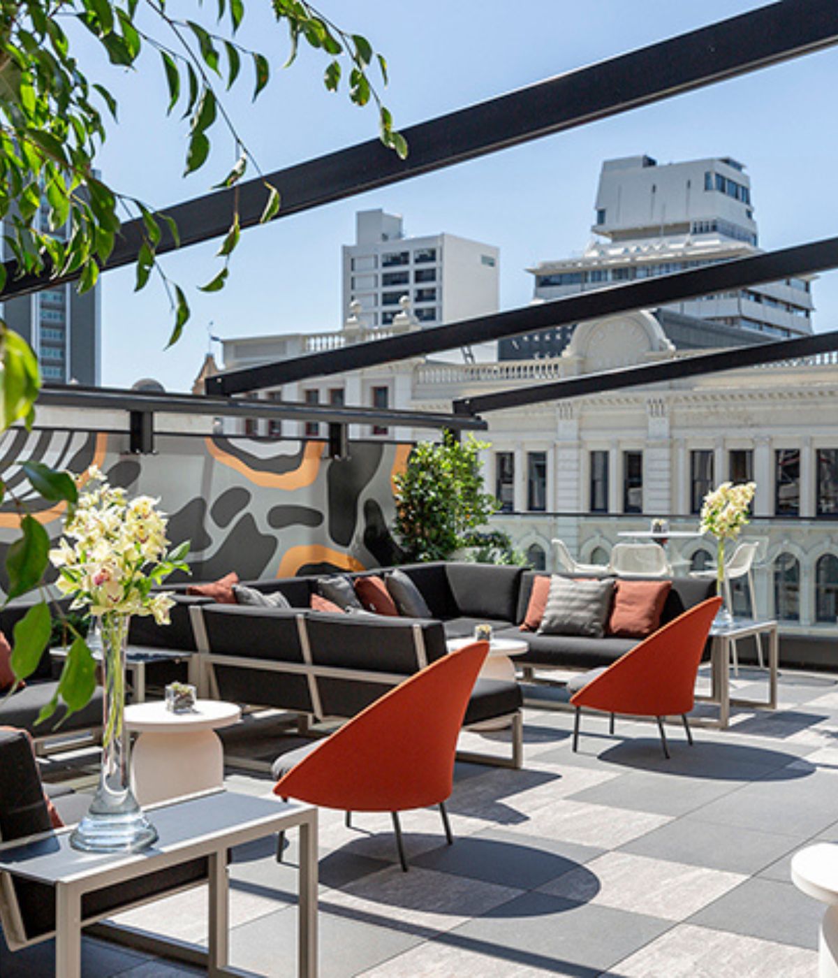 Rooftop terrace with dark sofas, orange chairs, geometric mural, and city buildings in background