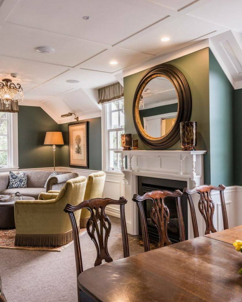 Green-walled lounge with white fireplace, chandelier, timber dining chairs and velvet armchairs