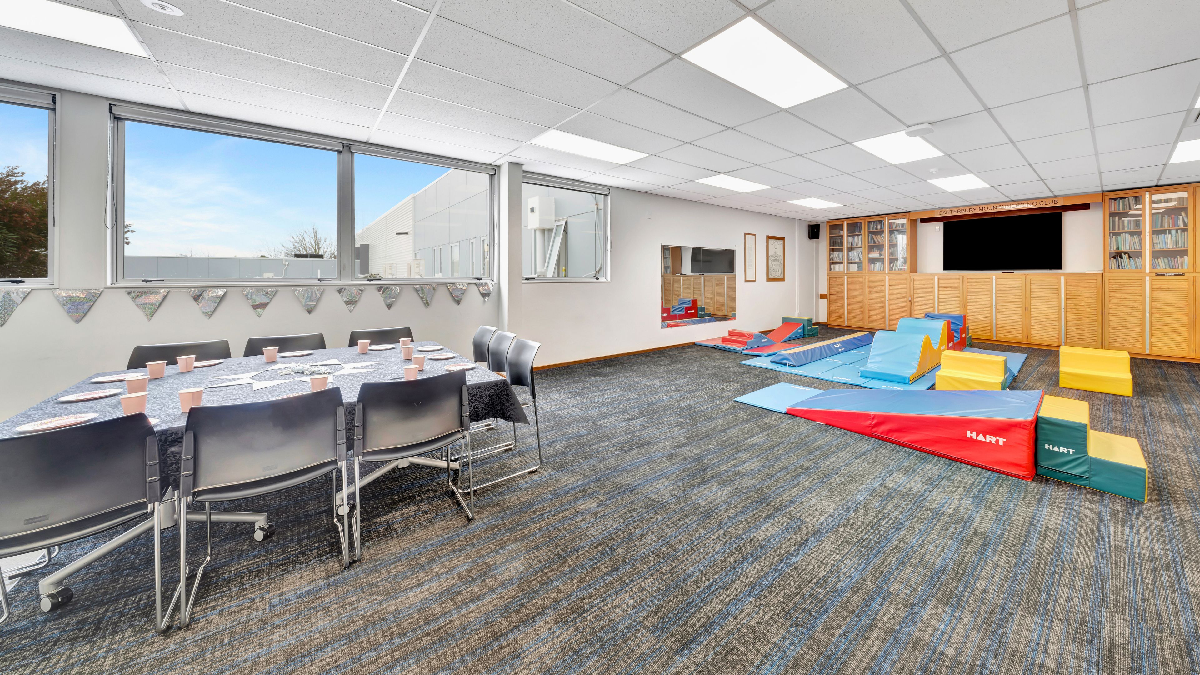 Carpeted activity room with round table, chairs and foam play equipment beside windows