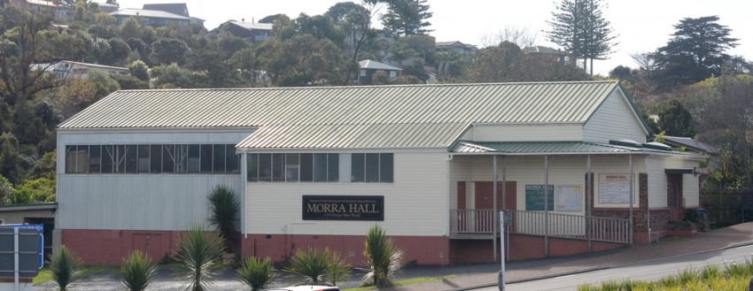 Low-pitched cream and red-brick community hall exterior with green corrugated roof, hillside behind