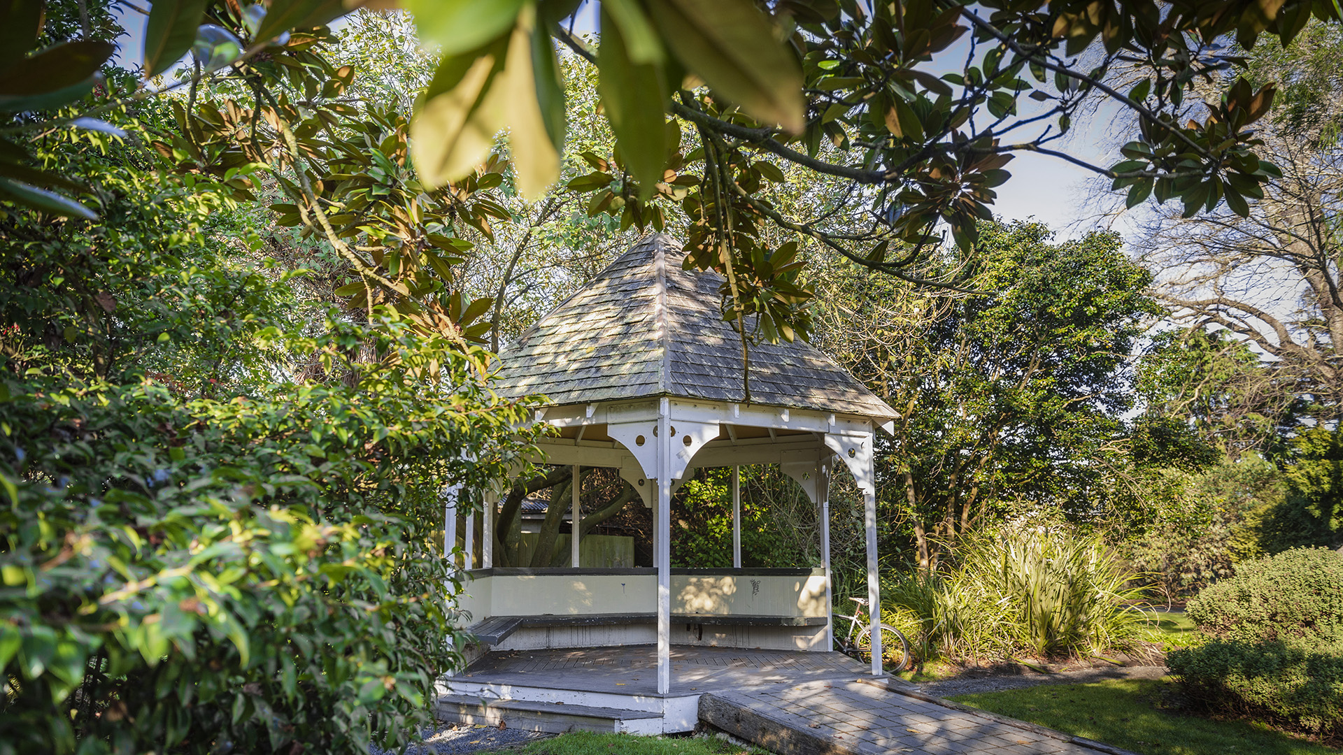 White wooden gazebo with shingled roof set among dense mature trees and garden
