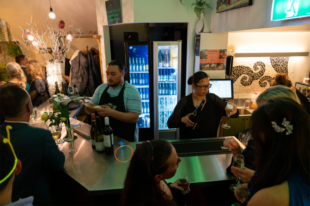 Bar service area with illuminated drinks fridge and bartender, decorated interior space with guests in background