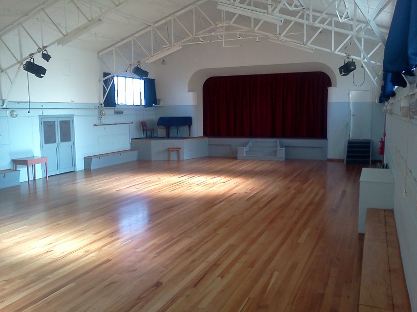 White-painted hall interior with timber floor, exposed roof trusses, and raised platform with red curtain