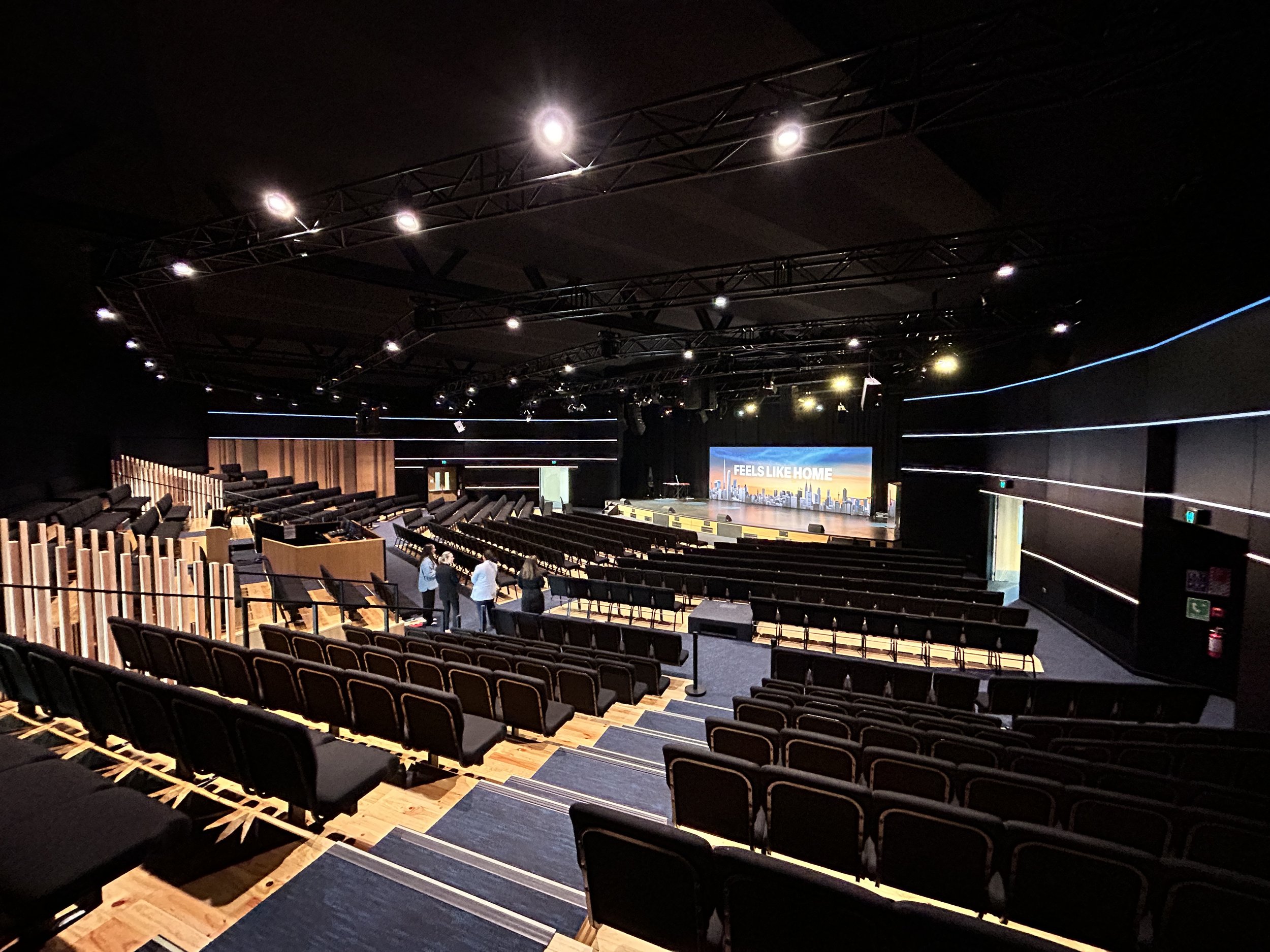 Large auditorium with tiered seating, raised stage with LED screen, and theatrical lighting rig